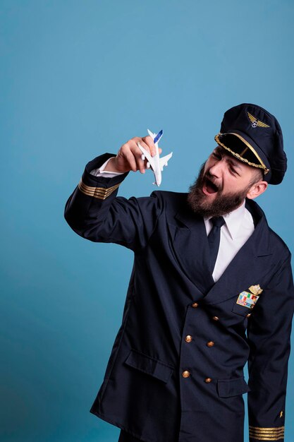 Photo smiling aviator in uniform holding airplane model, aviation academy captain playing with plane. aircraft crew person looking at commercial jet toy, side view studio medium shot