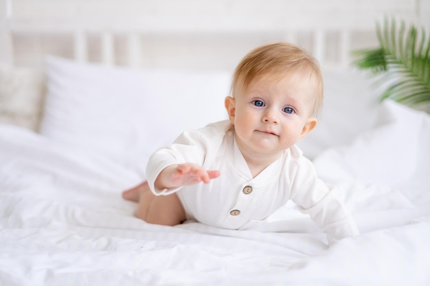 Smiling baby 6 months old blond boy crawls on a white bed in a bright bedroom after sleeping in the morning in a cotton bodysuit the concept of children's goods