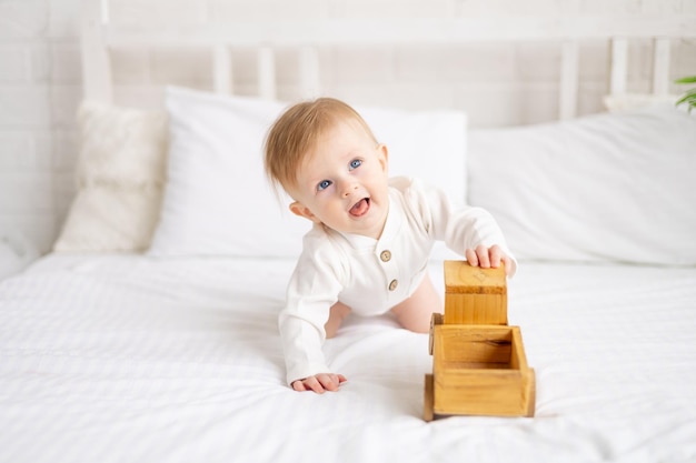 Улыбающийся малыш 6 месяцев блондин сидит на большой кровати в светлой спальне и играет с деревянной игрушечной машинкой в хлопковом комбинезоне концепция детских товаров