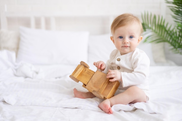 Smiling baby 6 months old blond boy is sitting on a large bed in a bright bedroom and playing with a wooden toy car in a cotton bodysuit the concept of children's goods