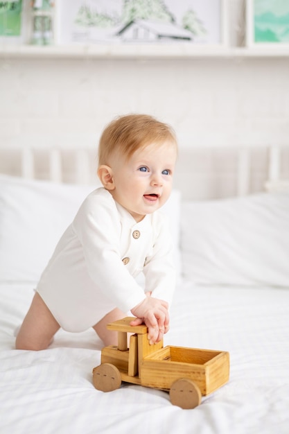 Smiling baby 6 months old blond boy is sitting on a large bed in a bright bedroom and playing with a wooden toy car in a cotton bodysuit the concept of children's goods