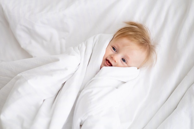 Smiling baby 6 months old blond boy lies on a white bed in a bright bedroom in a blanket after sleeping in the morning cotton bodysuit the concept of children's goods