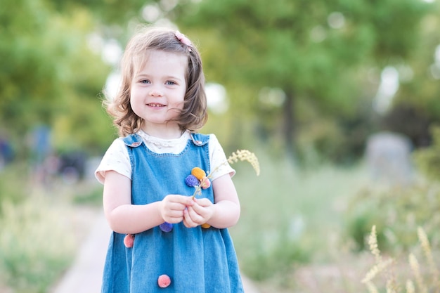 들판에 꽃을 들고 있는 트렌디한 데님 드레스를 입고 웃고 있는 아기 소녀
