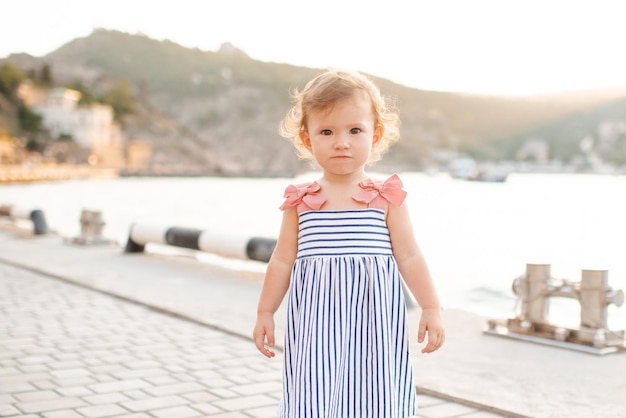 Smiling baby girl 1-2 year old wear striped dress and holding walk over wooden pier at sea shore outdoors. Summer season. Childhood. Looking at camera.