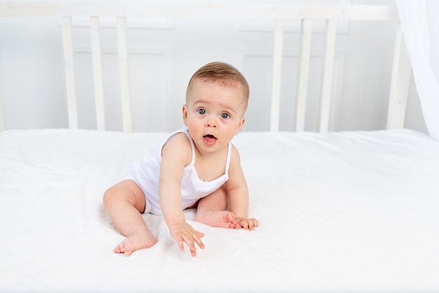 Smiling baby girl 8 months old sitting in a crib in a children's room in white clothes and l, baby's morning, baby products concept