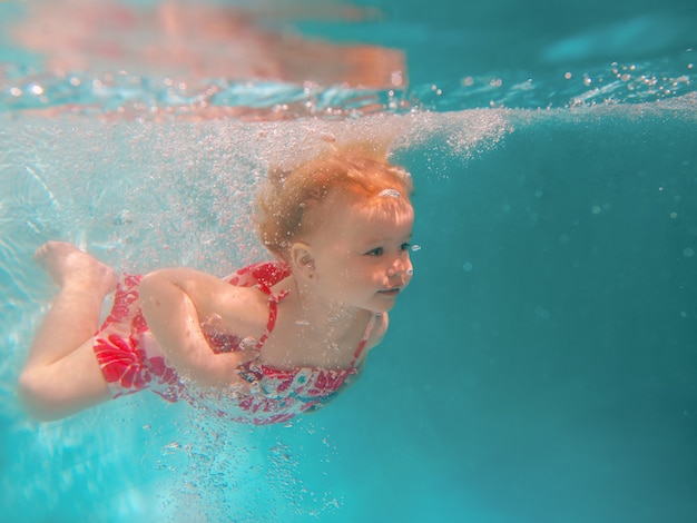 Smiling baby girl in cute modern dress diving underwater in blue swimming pool