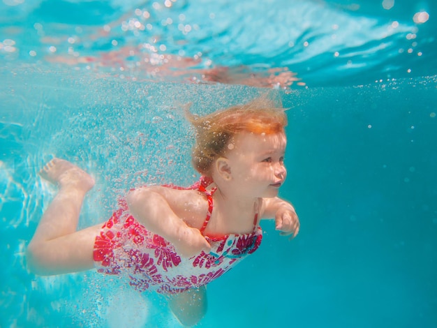 Smiling baby girl in cute modern dress diving underwater in blue swimming pool