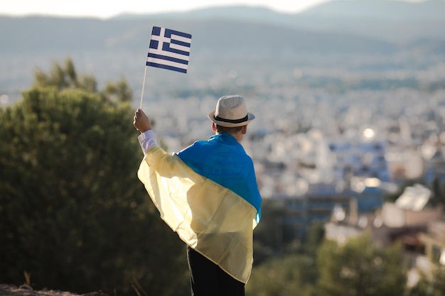 Smiling baby toddler boy holding a Greece and Ukrainian flag on city background Friendship country