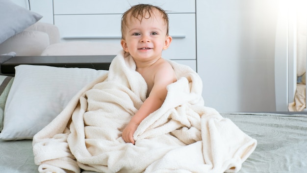 Photo smiling baby in white towel after having bath sitting on bed.