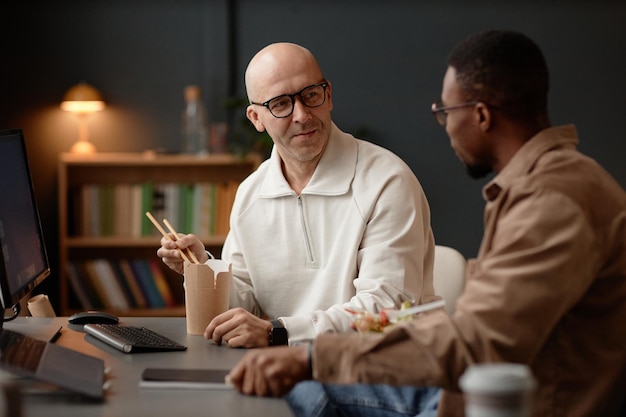 smiling bald man having lunch takeaways together with coworker at desk in office