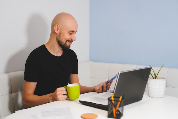 Smiling bald man with beard in black Tshirt looking to phone in his hand and holding cap in another