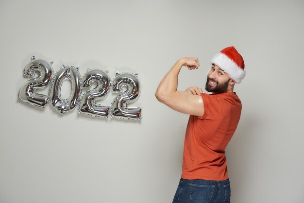 Foto un uomo calvo sorridente con la barba in un cappello da babbo natale mostra i bicipiti posteriori con un cerotto dopo la vaccinazione contro il coronavirus (covid-19) vicino a palloncini d'argento sventati a forma di 2022.