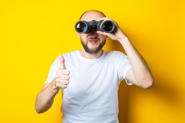 Smiling bald young man with a beard looks through binoculars and shows a thumb up