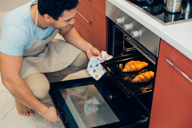 Smiling barefooted man in an apron squatting down by the cooker and taking a backing pan from the oven