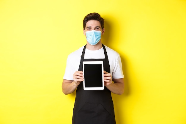 Smiling barista in medical mask showing tablet screen, standing over yellow wall