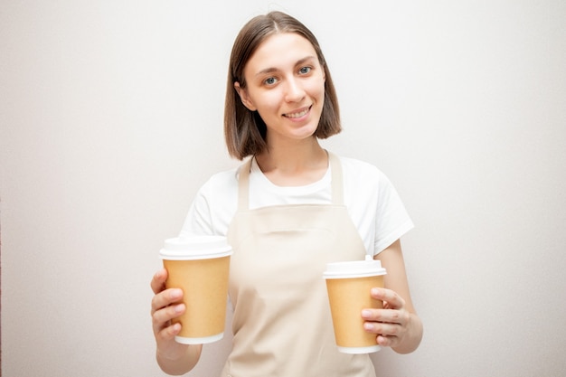 Smiling barista wearing apron holding two brown paper coffee cups. Woman in apron looking at camera and smiling.