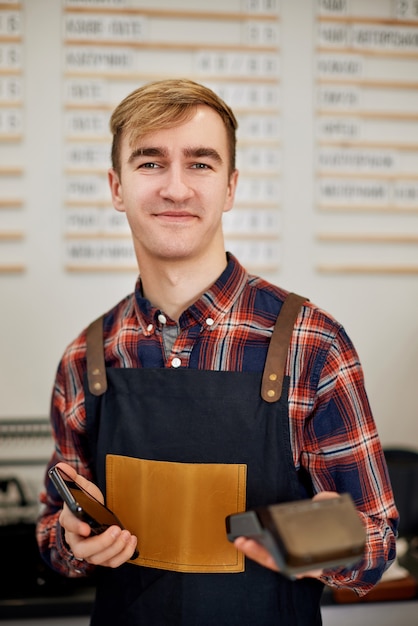 Smiling barman in apron hold modern bank payment terminal in cafe