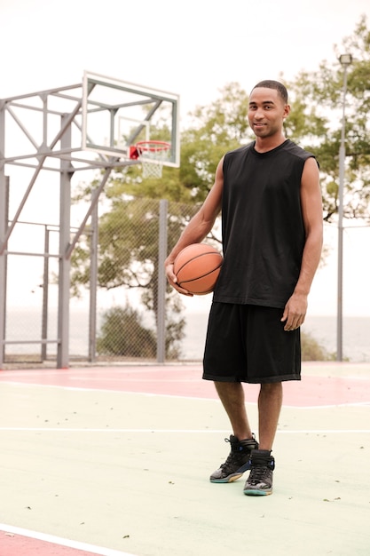 Smiling basketball player standing in the street with basketball hoop at wall