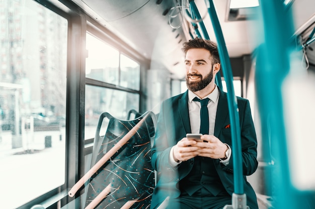 Smiling bearded Caucasian businessman in formal wear sitting in public transport and using smart phone.