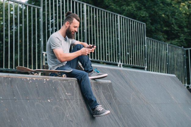 Smiling bearded hipster sitting on ramp and browsing smartphone.