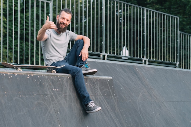 Smiling bearded hipster sitting on ramp and showing shaka sign.