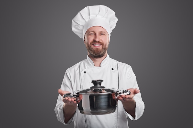 Smiling bearded male cook in toque and uniform with pot looking at camera on gray background