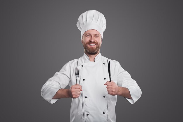 Smiling bearded male cook in uniform with toque holding fork and knife while looking at camera