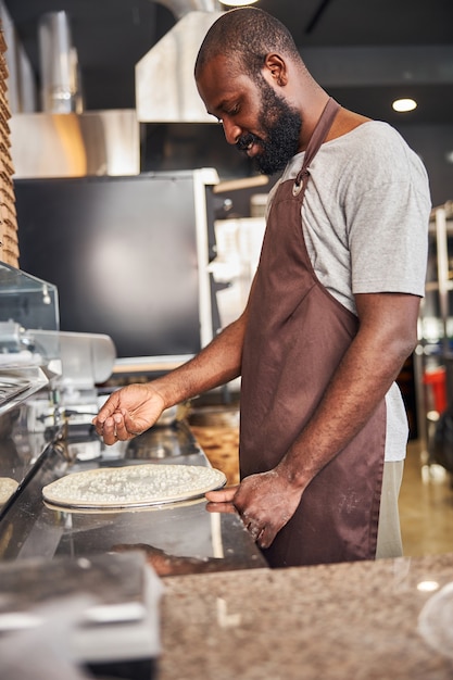 Photo smiling bearded man in apron is standing at table and putting grated cheese on dough