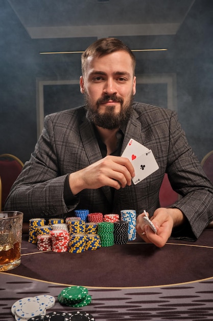 Smiling bearded man in a classic suit is playing poker sitting at the table at casino in a smoke. He is showing two aces and chips at the camera and realising that has won. Gambling for money. Games o