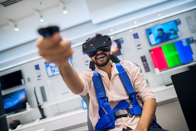 улыбающийся бородатый мужчина, держащий в руке беспроводной джойстик, тестирует очки виртуальной реальности и сидит в кресле с ремнями безопасности в техническом магазине