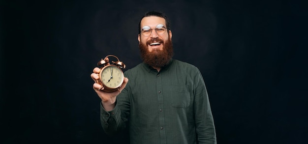 Smiling bearded man is holding an alarm clock while looking at the camera Studio shot over black background