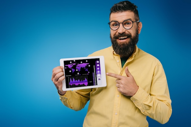 Smiling bearded man pointing at digital tablet with infographic isolated on blue