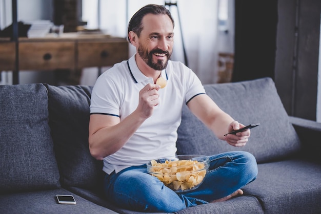 Smiling bearded man sitting on the sofa watching tv and eating chips at home