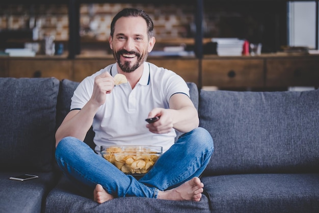 Smiling bearded man sitting on the sofa watching tv and eating chips at home