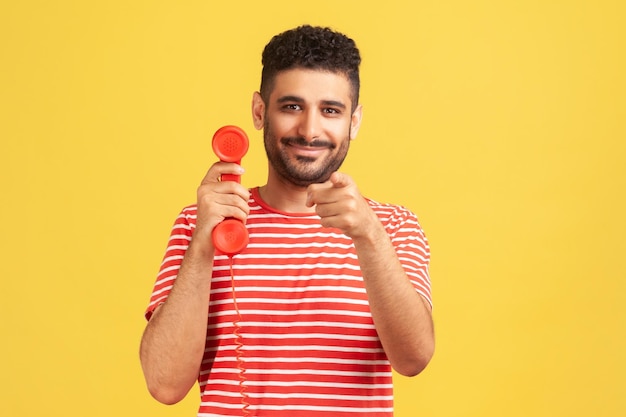 Улыбающийся бородатый мужчина в полосатой красной футболке, указывающий пальцем на вас, держащий в руках стационарный телефон, ожидающий вашего звонка. Внутренний студийный снимок изолирован на желтом фоне