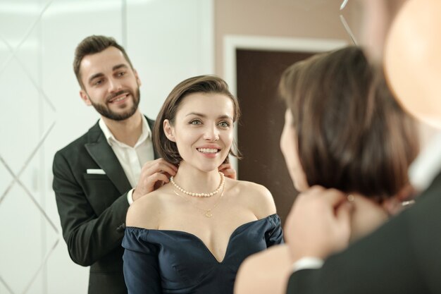 Smiling bearded man in suit wearing necklace to girlfriend in front of mirror while they getting dressed for dinner in restaurant