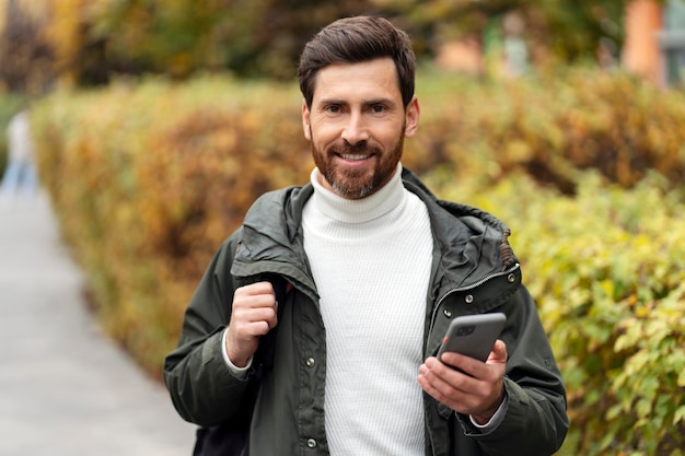 Smiling bearded man using mobile phone holding backpack Confident guy outdoors in the middle of the city