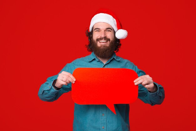 Smiling bearded man wearing christmas hat holds a speech bubble over red background