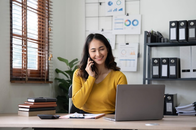 Smiling beautiful Asian businesswoman analyzing chart and graph showing changes on the market and holding smartphone at office