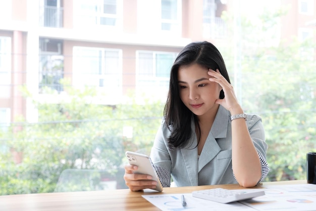 웃고 있는 아름다운 아시아 여성 사업가가 시장의 변화를 보여주는 차트와 그래프를 분석하고 사무실에서 스마트폰을 들고 있다