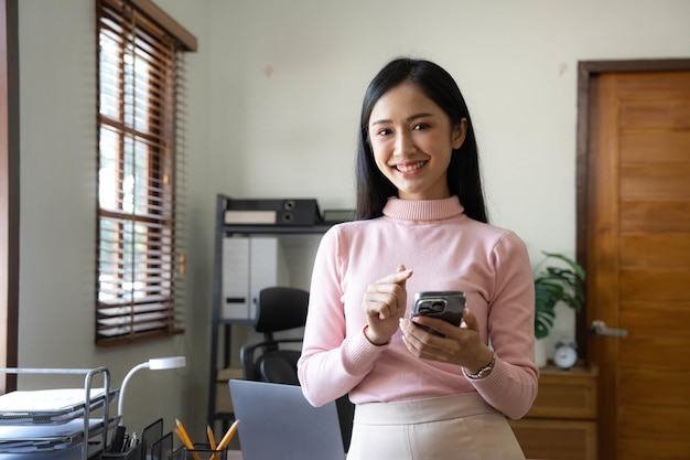 Smiling beautiful Asian businesswoman analyzing chart and graph showing changes on the market and holding smartphone at office