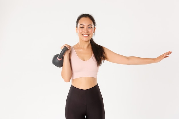 Smiling beautiful asian fitness girl, gym coach extend one hand and lift kettlebell, bodybuilding, gaining muscle strength, standing white background.