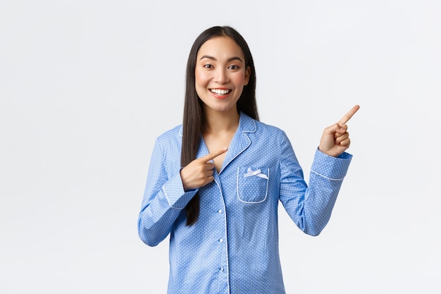 Smiling beautiful asian girl in blue pajamas making announcement, pointing fingers upper right corner to show big news or promo banner, recommend advertisement over white wall