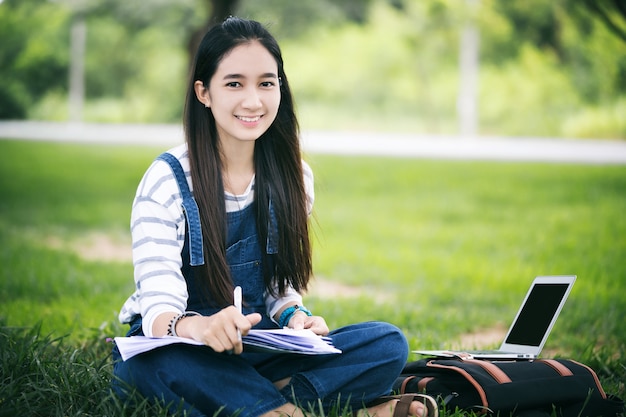 美しいアジアの女の子の読書の笑顔と、夏の公園の木の葉で働いてくつろげる時間