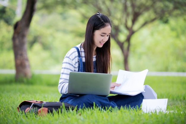 Il bello libro di lettura asiatico sorridente della ragazza e lavorare all'albero sul parco di estate per si rilassano il tempo