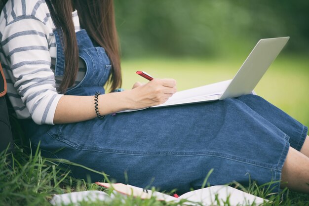 Smiling beautiful Asian girl reading book and working at tree on park in summer for relax time