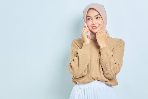 Bella donna musulmana asiatica sorridente in maglione marrone e hijab che parla sul telefono cellulare mentre guarda da parte isolato su sfondo bianco concetto di stile di vita religioso della gente