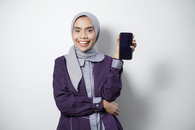 Smiling beautiful Asian woman in casual shirt and hijab holding a mobile phone with a blank screen isolated by white background