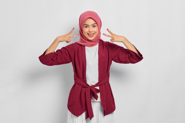 Smiling beautiful Asian woman in casual shirt showing peace sign gesture and looking at camera isolated over white background
