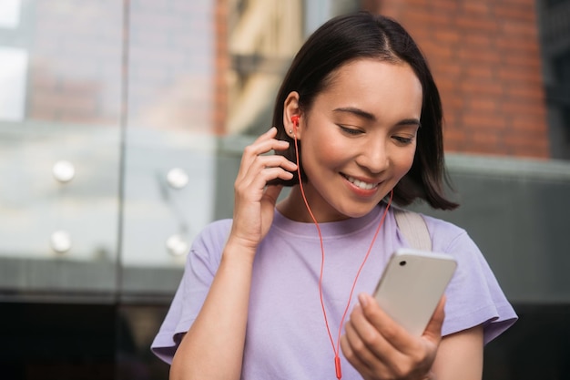 Smiling beautiful asian woman holding mobile phone listening music, watching video on the street